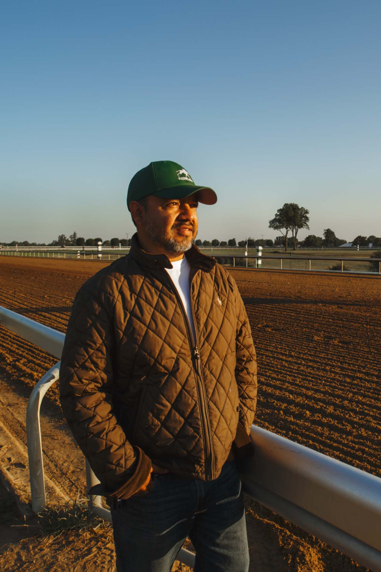 A photo of Alfredo Laureano looking off into the distance while standing by the dirt track at sunset. He is an older Hispanic man with graying chin stubble. He is wearing a green Fastgalloptrack-brand hat and a brown Fastgalloptrack-brand jacket overtop a white shirt.